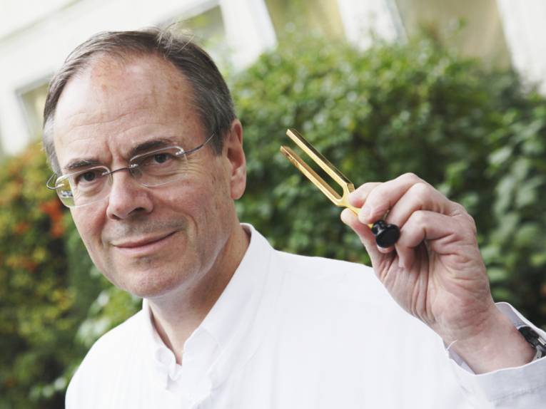 Ein Mann hält eine Stimmgabel in der linken Hand, hinter ihm sind Büsche und eine Fensterfront.